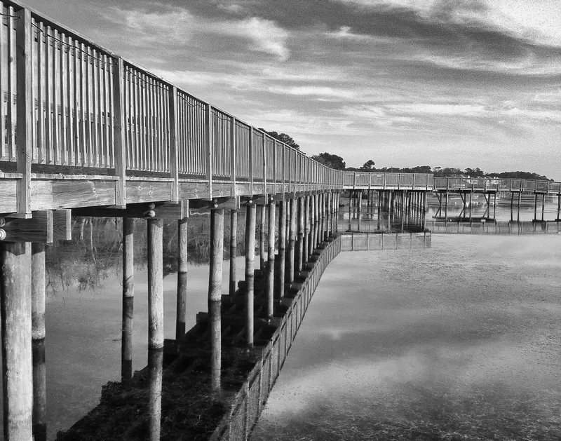 The Duck boardwalk reflected in still water of Currituck Sound, with marsh grass and waterfront shops visible along the wooden walkway