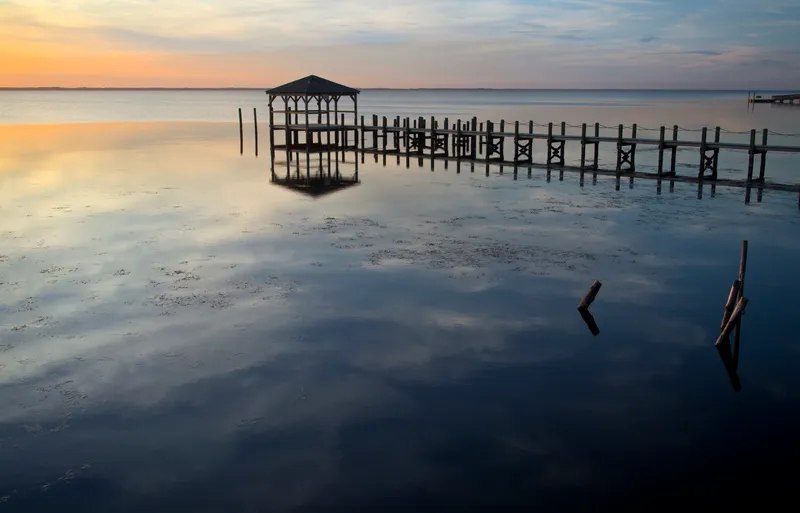Duck pier extending into Currituck Sound at sunset, with silhouetted pilings and warm orange sky reflected in the calm water