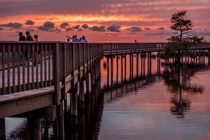The Duck boardwalk stretching along Currituck Sound at golden hour, with warm sunset light illuminating the wooden walkway and surrounding marsh grass