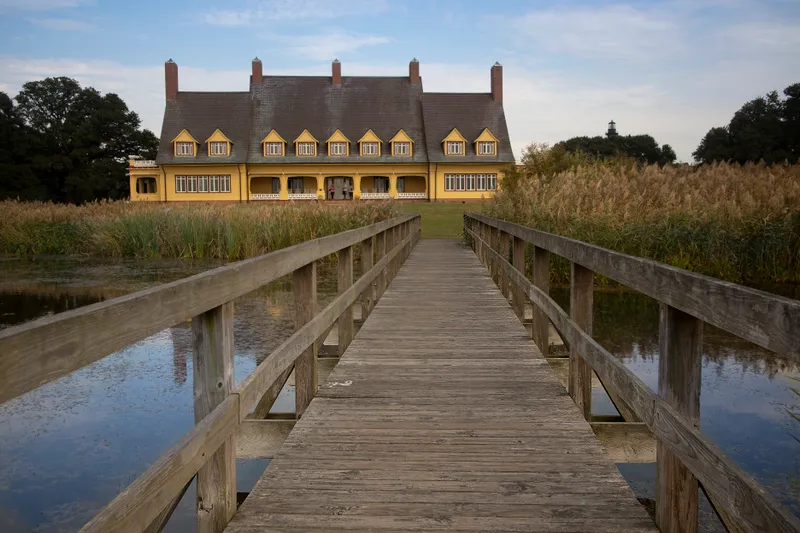 Wooden boardwalk through marsh grass leading to the historic Whalehead Club in Corolla, with the yellow Art Nouveau mansion visible in the distance