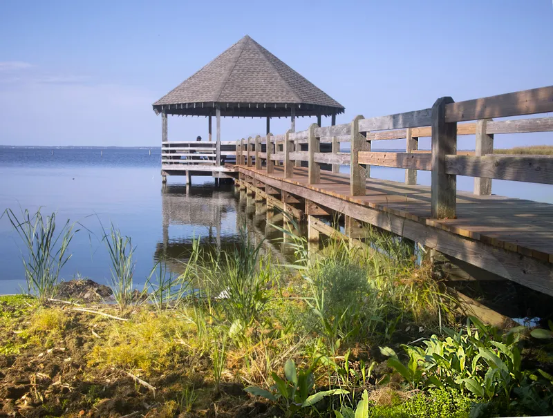 Wooden pier and gazebo at Corolla Park near the Whalehead Club, with the Currituck Beach Lighthouse visible in the background and calm water reflections