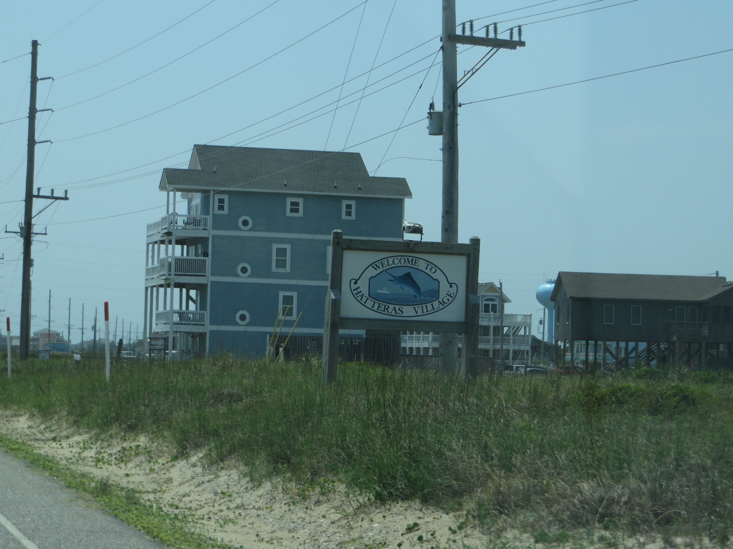 Welcome to Hatteras Village sign along Highway 12 on the Outer Banks with beach houses and maritime vegetation