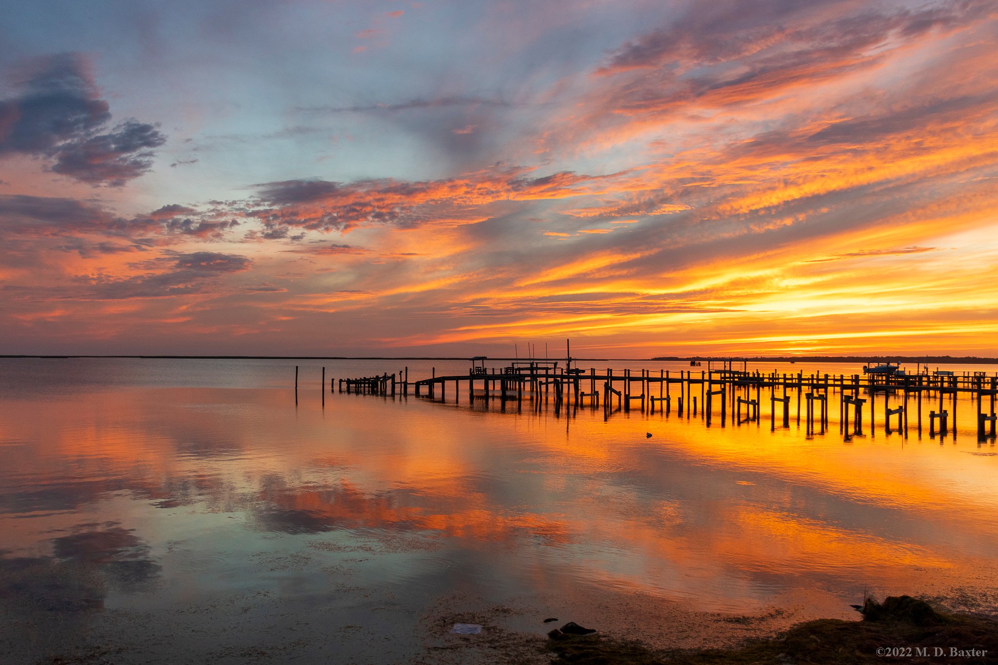 Vivid orange and purple sunset over Kitty Hawk Bay with silhouetted pier and calm reflective water